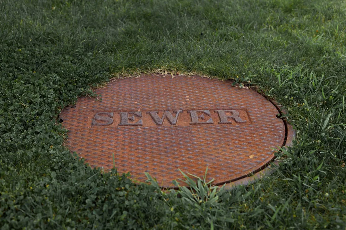 Close-up of a rusty sewer manhole cover surrounded by grass, highlighting potential sewer issues.
