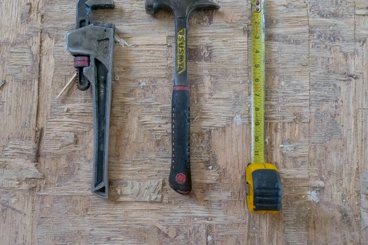 Close-up of a hammer, wrench, and measuring tape on a wooden surface, tools useful for water heater repair.
